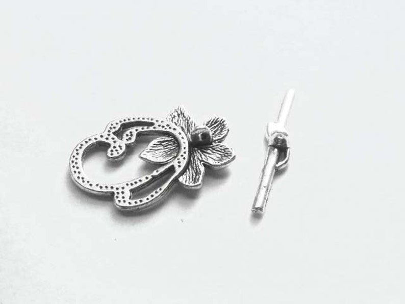 May include: Silver-toned metal clasp with a floral design. The clasp is made of two pieces, one with a decorative floral design and the other with a simple bar.
