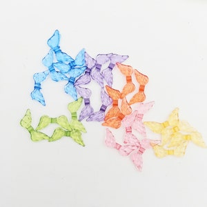 May include: Assortment of colorful plastic angel wings in various shades of blue, purple, orange, pink, and yellow. The wings are arranged in a loose pile on a white background.