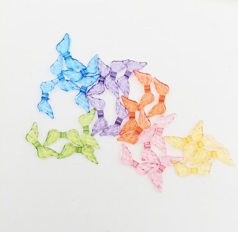 May include: Assortment of colorful plastic angel wings in various shades of blue, purple, orange, pink, and yellow. The wings are arranged in a loose pile on a white background.