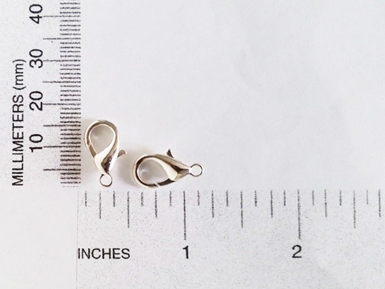 May include: Two silver-toned lobster clasps, each measuring approximately 1 inch in length, are shown against a white background with a ruler for scale.