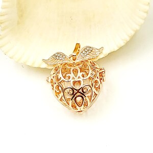May include: A gold-colored heart-shaped pendant with intricate filigree detailing. The pendant features small wings at the top and is displayed on a white background. The pendant is likely a piece of jewelry.