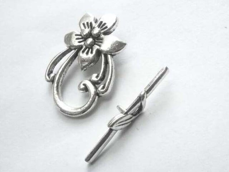 May include: Silver-toned metal toggle clasp with a floral design. The clasp features a decorative flower with a swirl design and a separate, straight bar with a leaf design.