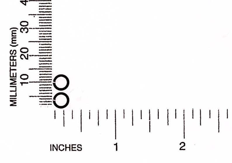 May include: Two small, black, circular objects are positioned near the 10 mm mark on a ruler. The ruler displays both inches and millimeters, with the inch markings below the millimeter markings.