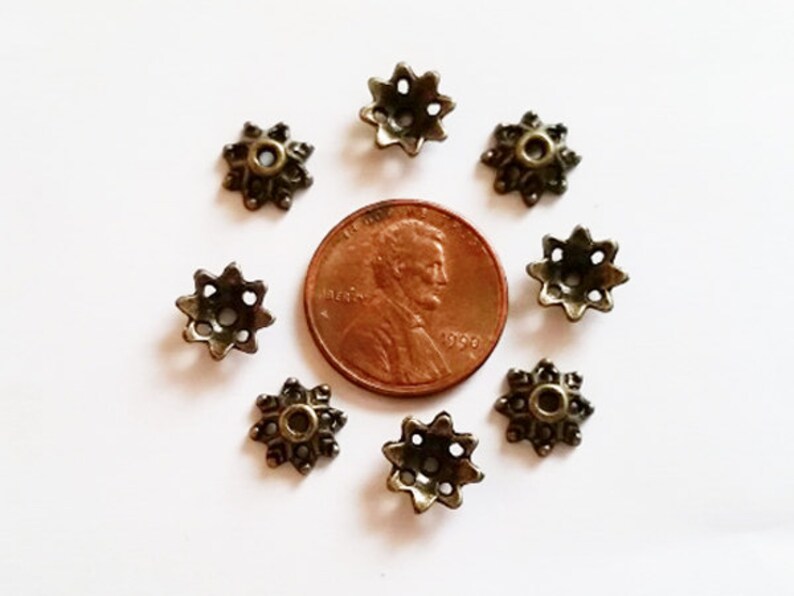 May include: Eight antique gold-toned metal star-shaped embellishments with a hole in the center. The embellishments are arranged in a circle around a US penny.