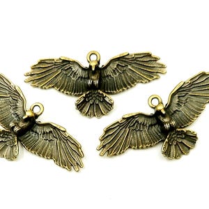 Puede incluir: Tres colgantes de metal color bronce con forma de águila y alas extendidas. Cada colgante tiene un lazo en la parte superior para sujetarlo a joyas u otras manualidades. Las águilas están detalladas con patrones de plumas y alas.