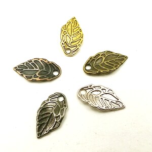 May include: Four metal leaf charms in different finishes: gold, bronze, silver, and black. Each charm has a delicate, intricate design and a small hole for attaching to jewelry or crafts.