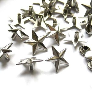 Puede incluir: Una colecci&oacute;n de tachuelas de metal en forma de estrella de color plateado con un centro puntiagudo. Las tachuelas tienen un agujero en el centro para sujetarlas a tela u otros materiales.