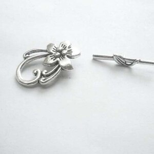 May include: Silver-toned metal flower charm with a decorative swirl design and a matching silver-toned metal bar with a leaf design.