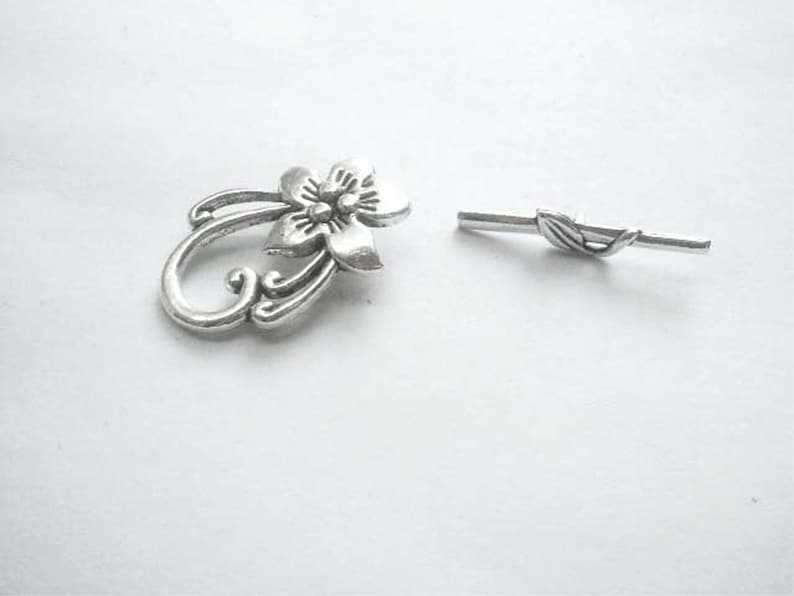 May include: Silver-toned metal flower charm with a decorative swirl design and a matching silver-toned metal bar with a leaf design.