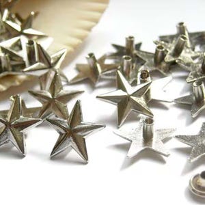 Puede incluir: Una gran cantidad de tachuelas de metal en forma de estrella de color plateado. Las tachuelas tienen una punta puntiaguda y una parte posterior plana con un agujero en el centro. Las tachuelas est&aacute;n dispersas sobre una superficie blanca.