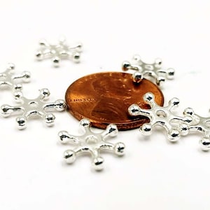 May include: Silver snowflake charms, scattered on a white surface, with a copper penny in the center.