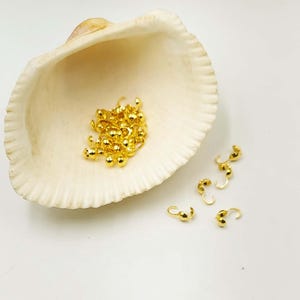 Può includere: Una collezione di piccoli elementi di gioielleria dorati e lucenti, tra cui copri perline a crimpare e chiusure a forma di gancio. Gli elementi sono esposti in una conchiglia bianca su sfondo bianco, creando un contrasto di trame e colori.