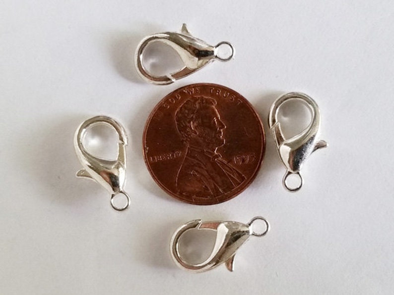 May include: Four silver-toned lobster claw clasps, each with a small loop, are arranged around a US penny. The penny is dated 1995 and has the inscription "In God We Trust" and "Liberty".