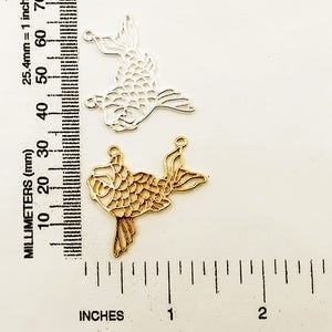 May include: Two metal fish charms, one gold and one silver, with a detailed scale pattern. The charms are approximately 2 inches long and are shown next to a ruler.
