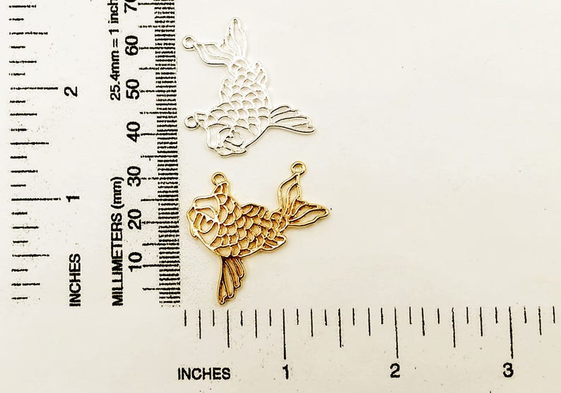 May include: Two metal fish charms, one gold and one silver, with a detailed scale pattern. The charms are approximately 2 inches long and are shown next to a ruler.