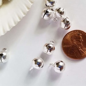 May include: Silver jingle bells with a shiny finish. The bells are scattered on a white surface next to a US penny.