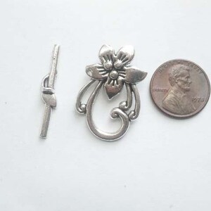 May include: Silver tone metal clasp with a floral design. The clasp is about the size of a US penny.