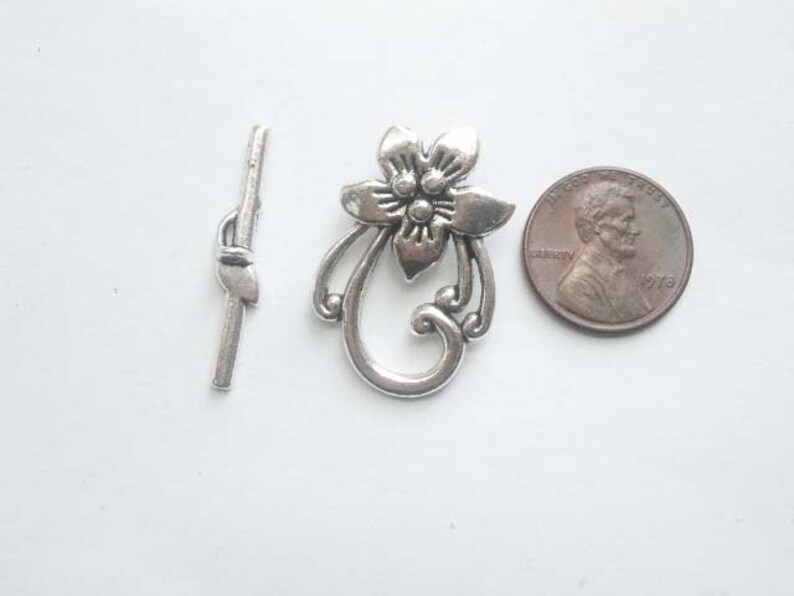 May include: Silver tone metal clasp with a floral design. The clasp is about the size of a US penny.