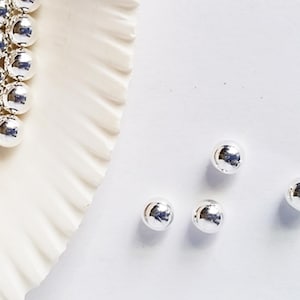 May include: Four silver metal beads on a white surface. The beads are round and shiny.
