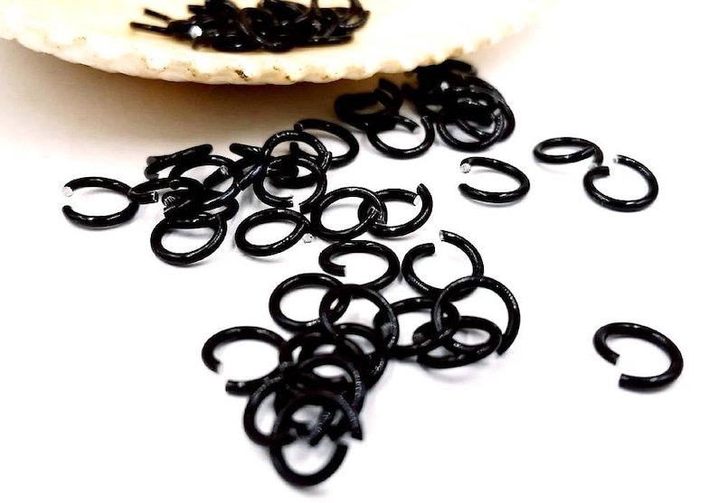 May include: A collection of small, black metal jump rings scattered on a white surface. Some rings are open, while others are closed. A few jump rings are in a white, shell-shaped dish.