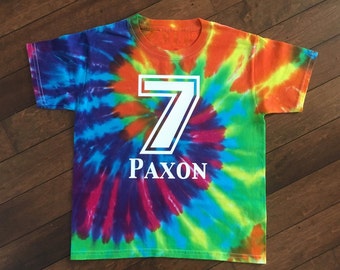 May include: A colorful tie-dye t-shirt with a white number 7 and the name "PAXON" printed on the front. The shirt features a spiral design with vibrant hues of blue, green, orange, and purple. This shirt is perfect for a birthday.