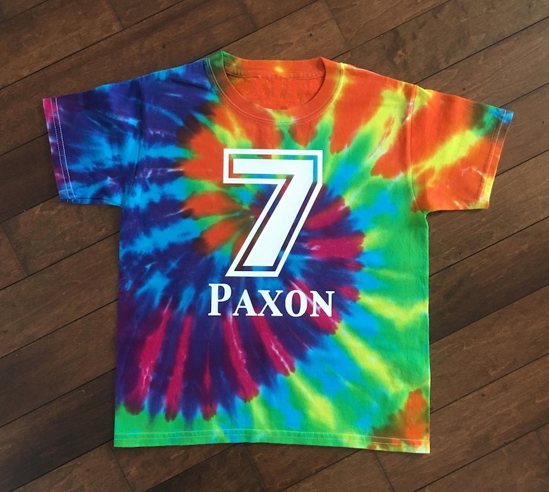 Pode incluir: Uma t-shirt tie-dye colorida com o n&uacute;mero 7 branco e o nome "PAXON" impresso na frente. A t-shirt apresenta um design em espiral com tons vibrantes de azul, verde, laranja e roxo. Perfeita para um anivers&aacute;rio.