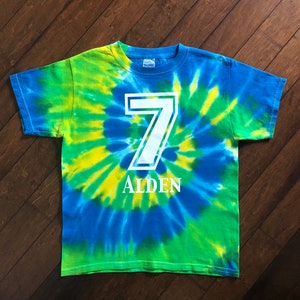 Pode incluir: Uma t-shirt tie-dye em tons de azul, verde e amarelo. O n&uacute;mero 7 &eacute; impresso em branco, com o nome "ALDEN" abaixo. A t-shirt &eacute; da marca Gildan.