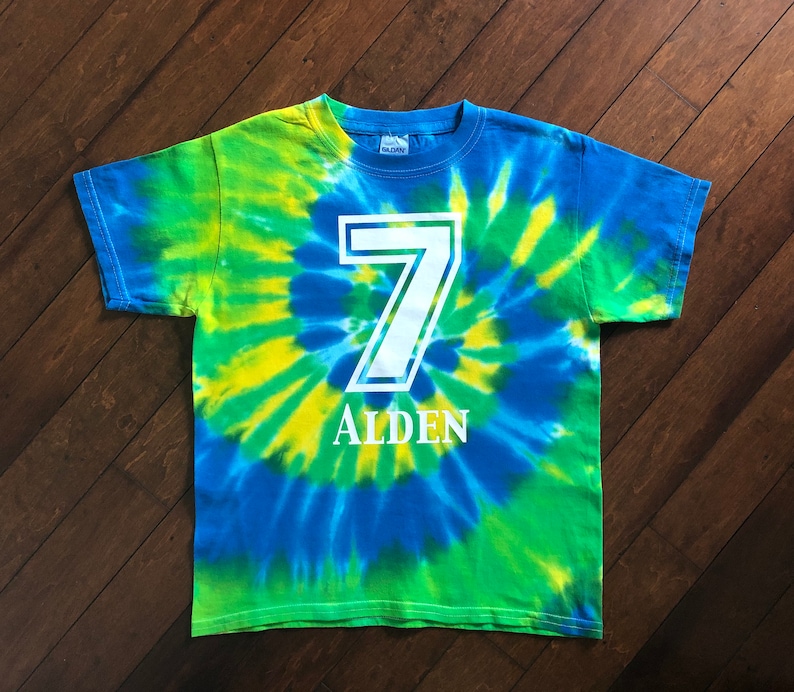 Pode incluir: Uma t-shirt tie-dye em tons de azul, verde e amarelo. O n&uacute;mero 7 &eacute; impresso em branco, com o nome "ALDEN" abaixo. A t-shirt &eacute; da marca Gildan.