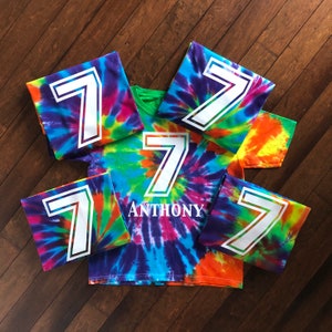 Pode incluir: Camisetas tie-dye com um grande n&uacute;mero 7 branco. As camisas apresentam uma mistura de cores vibrantes, incluindo azul, roxo, verde, laranja e amarelo. Uma camisa tem o nome "Anthony". As camisas est&atilde;o dispostas sobre uma superf&iacute;cie de madeira.