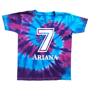 Pode incluir: Uma camiseta tie-dye em tons de azul e roxo, com um grande n&uacute;mero 7 branco e o nome "ARIANA" em letras brancas. A camiseta &eacute; de manga curta e gola redonda.