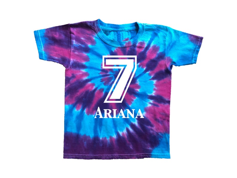 Pode incluir: Uma camiseta tie-dye em tons de azul e roxo, com um grande n&uacute;mero 7 branco e o nome "ARIANA" em letras brancas. A camiseta &eacute; de manga curta e gola redonda.