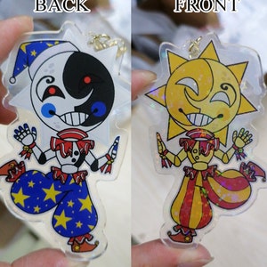 May include: Two-sided acrylic charm featuring a cartoon character with a jester hat and a star-shaped head. The front side is yellow with a smiling sun face, and the back side is black and white with a crescent moon face. Both sides have a red and white jester collar and red and white pants with yellow stars.