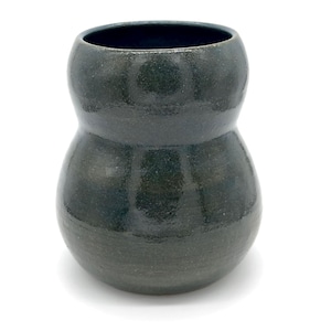 Pode incluir: Um vaso de cerâmica verde escuro com uma forma de ampulheta única. O vaso tem uma abertura larga e uma superfície texturizada, dando-lhe uma aparência artesanal. A cor verde escura adiciona um toque de elegância.