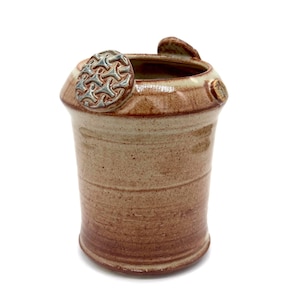 Pode incluir: Um recipiente de cerâmica feito à mão com formato cilíndrico. A cerâmica é principalmente castanha com detalhes castanhos e um emblema circular decorativo na lateral. A borda tem uma cor bege clara. O recipiente tem uma estética rústica e terrosa.