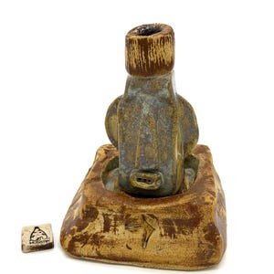 May include: A handcrafted ceramic incense holder with a unique, abstract design. The holder features a cylindrical top and a base with a textured, earthy appearance. The color palette includes shades of brown, tan, and blue-grey. A small, square tile with text is also present.