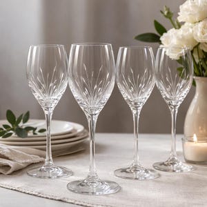 May include: Four clear crystal wine glasses with etched floral patterns. The glasses have long stems and round bases. A white vase with white flowers and a lit candle are in the background.