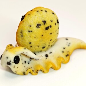 May include: A white and black cookie-themed snail figurine with a yellow shell and black chocolate chips. The snail is made of polymer clay and has a whimsical, cartoonish style.