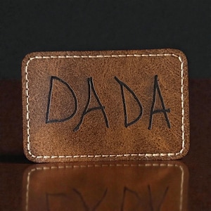May include: A rectangular brown leather patch with the word "DADA" in black, handwritten-style letters. The patch has a white stitched border. The item is likely a personalized gift or accessory.