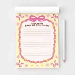 May include: A notepad with a light yellow background and a pink border. The notepad has the text "BUSY DOING SMALL BIZ GIRLIE THINGS" in the center. Pink bows and flowers decorate the edges. A white pencil is on the right.