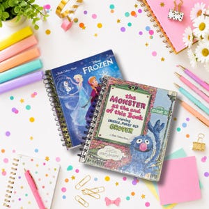 May include: Two spiral-bound notebooks featuring classic children's book covers. One notebook displays the Disney Frozen cover with Elsa and Olaf, while the other features The Monster at the End of This Book with Grover from Sesame Street.