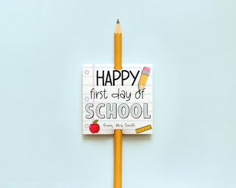 May include: A yellow pencil inserted through a square paper tag featuring a lined notebook pattern. The tag displays the text "HAPPY first day of SCHOOL" and "from, Mrs. Smith" with illustrations of a pencil, a red apple, and a yellow ruler.
