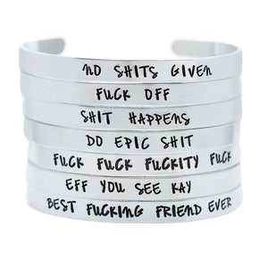 May include: A stack of silver metal cuff bracelets, each inscribed with a different phrase in black. The phrases include "NO SHITS GIVEN," "FUCK OFF," and "BEST FUCKING FRIEND EVER." The bracelets are displayed against a white backdrop.