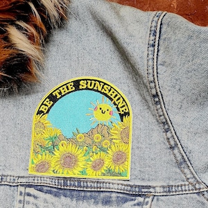 May include: A yellow and black embroidered patch with a smiling sun and sunflowers. The patch says "Be the Sunshine" in a black circle.