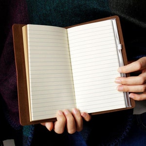 May include: Open brown leather journal with lined pages. The journal is held open, revealing the blank pages ready for writing. A clear plastic pen holder is attached to the spine. The journal is a classic and practical item for note-taking or journaling.