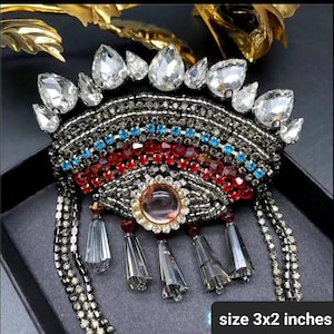 May include: A large, colorful brooch with a rainbow design made of beads and rhinestones. The brooch has a central eye-shaped design with a clear crystal in the center. The brooch is 3 inches by 2 inches.