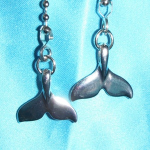 May include: A pair of silver-colored earrings, each featuring a whale tail design. The earrings hang from a small chain of silver beads. The background is a solid light blue fabric.