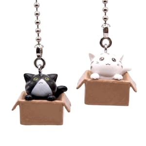 May include: Two cat-themed ceiling fan pull chains. One chain features a black and white cat in a brown cardboard box. The other chain has a white cat with gray markings in a brown cardboard box. Both chains have a silver-colored ball chain.