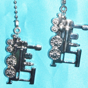 May include: Two silver-colored projector charms hanging from a chain. The charms are detailed with wheels and other mechanical elements, suggesting a vintage or retro theme. The background is a solid light blue.