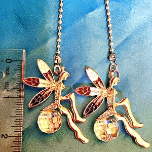 May include: Two decorative dragonfly-shaped ceiling fan pulls. Each dragonfly has gold-colored metal wings with brown, white, and black accents. A clear, faceted crystal ball hangs below each dragonfly. The pulls are approximately 1.5 inches long.