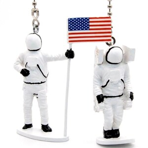 May include: Two white astronaut figurines with black accents, one holding a pole with a miniature American flag. Each figure is attached to a silver metal ball chain connector at the top, featuring detailed space suits and helmets on white bases.